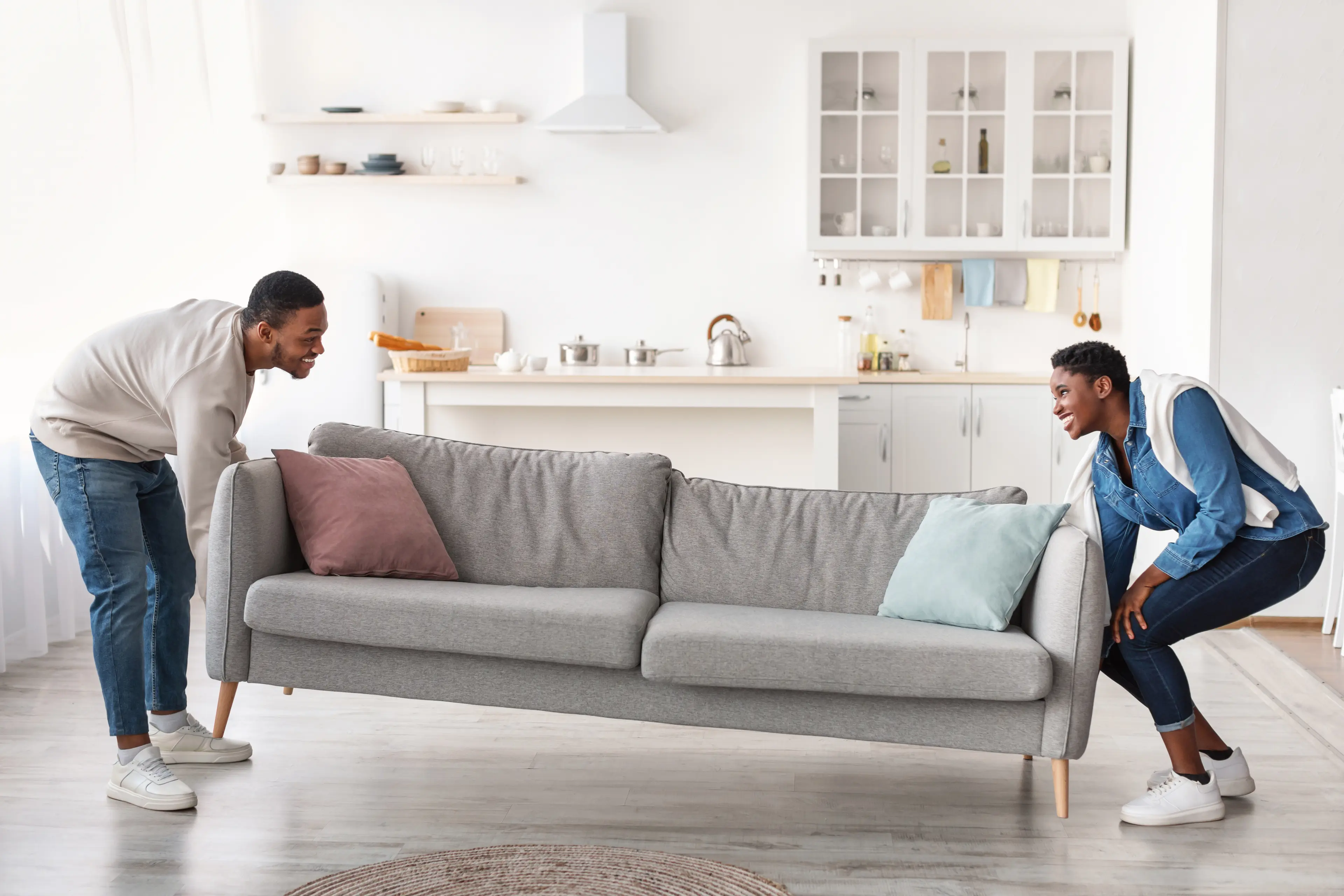 A smiling couple moves a sofa into a spacious, bright room. They look excited as they set up their new home, capturing the joy of starting fresh and creating a comfortable living space together. This shows how Acima Leasing can help customers get the items they need.